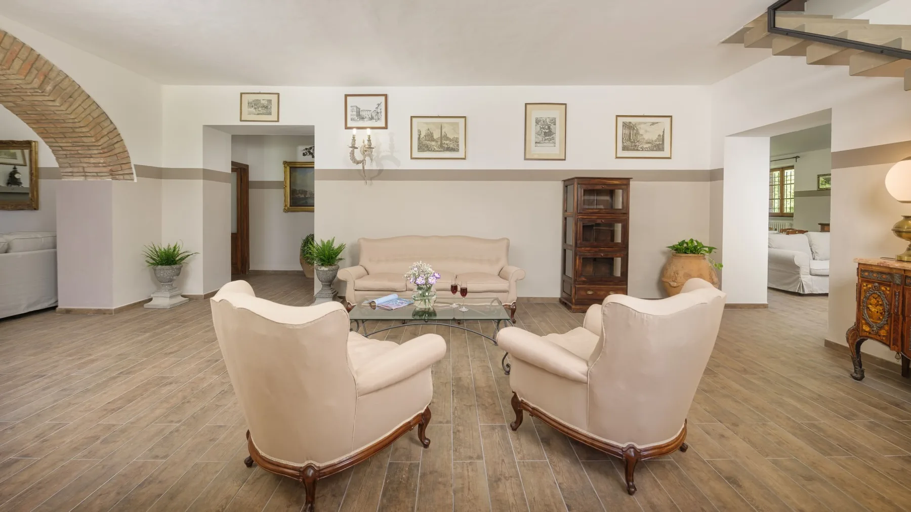 Una sala luminosa tiene dos sillones color crema, un sofá, cuadros enmarcados y una estantería de madera.