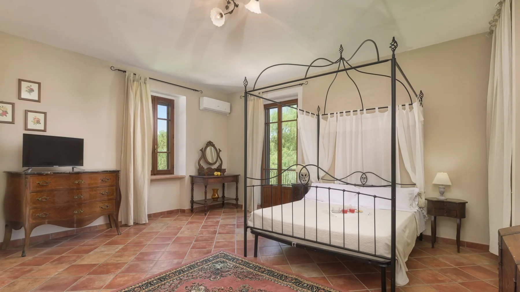 Un dormitorio con cama con dosel de hierro negro, cortinas blancas, suelo de baldosas de terracota y muebles de madera.