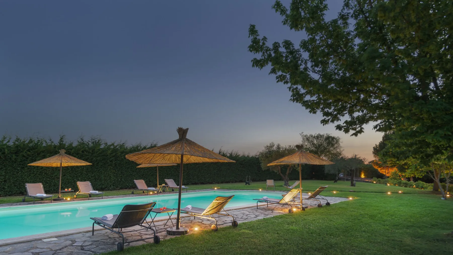 Una piscina al aire libre brilla al anochecer, con tumbonas, sombrillas y un amplio césped.