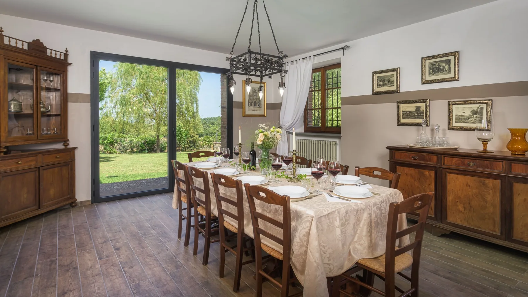 Un comedor con una mesa larga puesta, sillas de madera, cuadros y grandes ventanas con vistas al jardín.