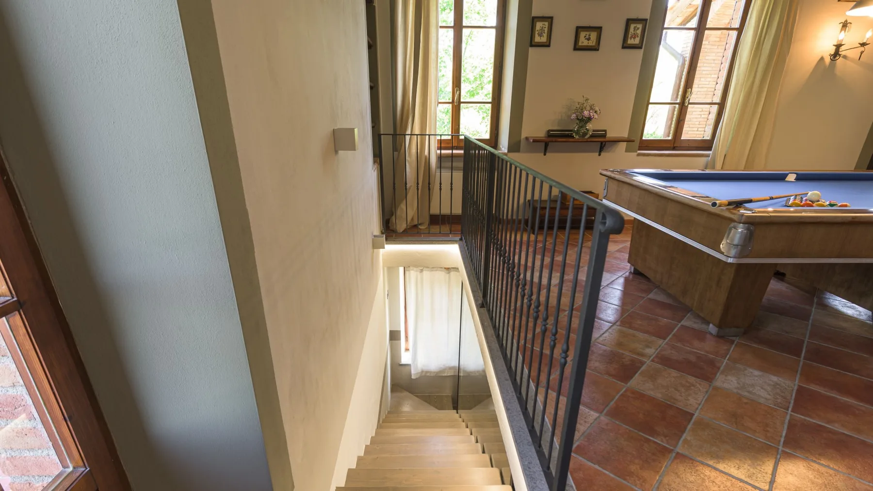 Una escalera con barandales negros da a una sala de juegos con piso de baldosas, mesa de billar y ventanas altas.