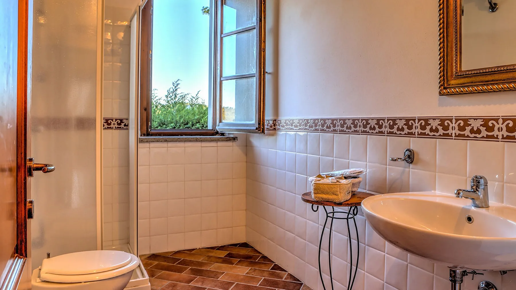 Un baño alicatado cuenta con un inodoro blanco, un lavabo amplio y una ventana abierta con vistas a la vegetación.