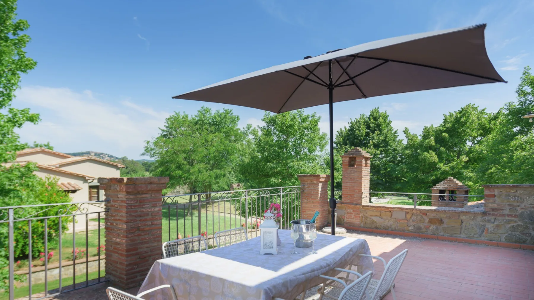 Un patio de ladrillo tiene una sombrilla grande que da sombra a una mesa y sillas junto a una barandilla metálica.