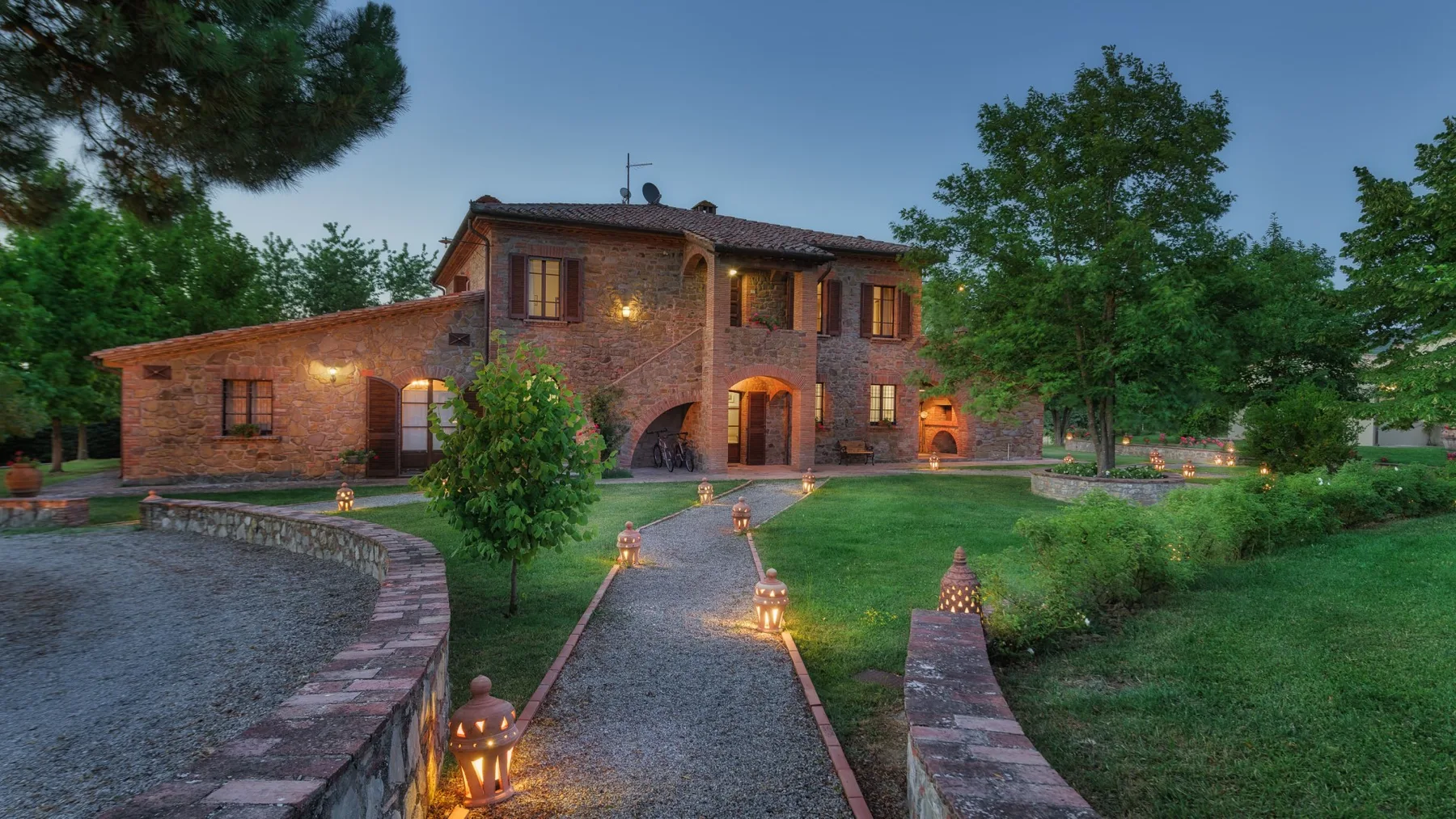 Una casa de ladrillo de dos plantas junto a una entrada de grava, con senderos de jardín y pequeñas luces al anochecer.