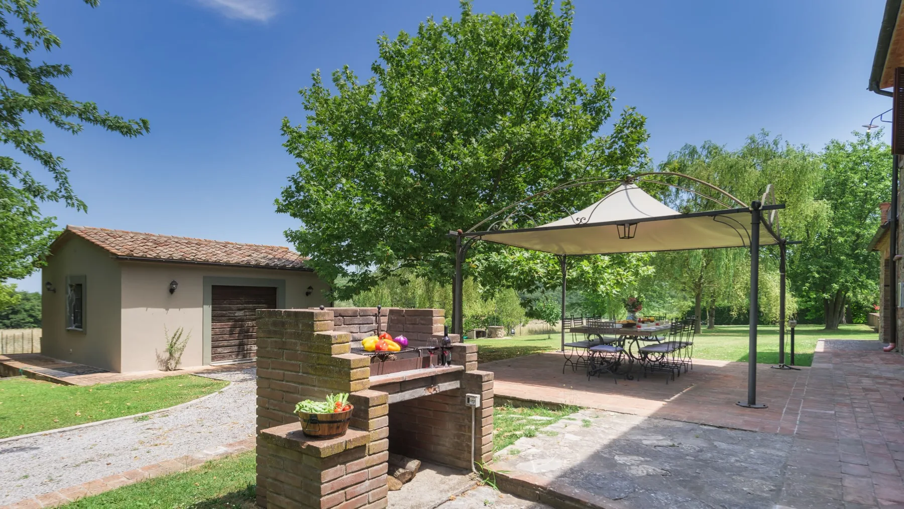 Una barbacoa exterior de ladrillo está junto a un patio sombreado con toldo, mesa de comedor y jardín frondoso.