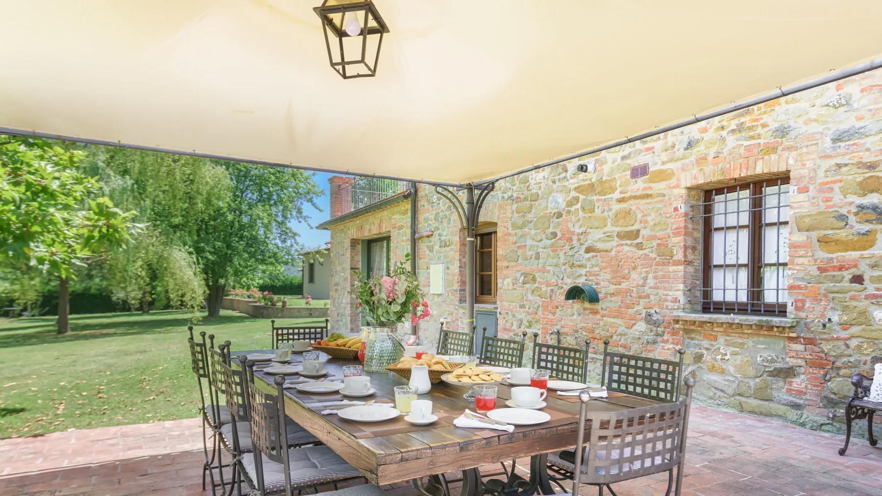 Una zona de comedor exterior a la sombra tiene una larga mesa de madera con platos junto a un muro de piedra.