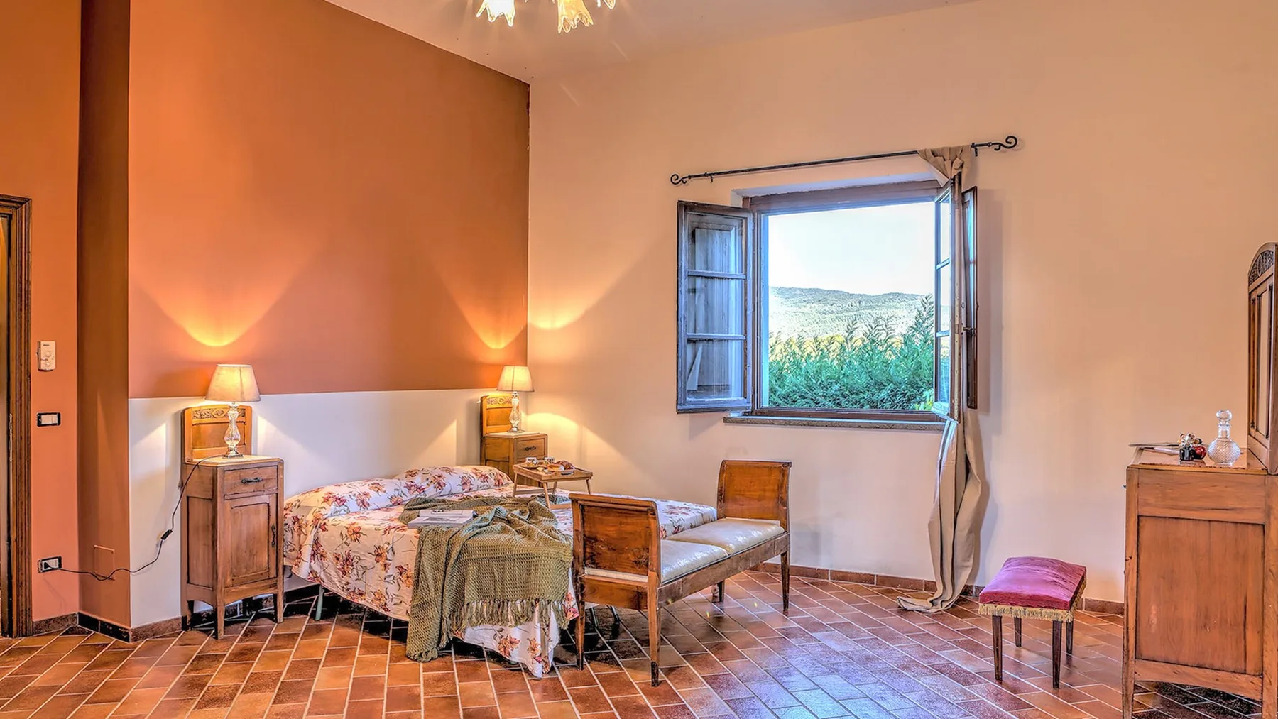 Un dormitorio de tonos cálidos tiene una cama con flores, muebles de madera y una ventana abierta con vistas a la vegetación.