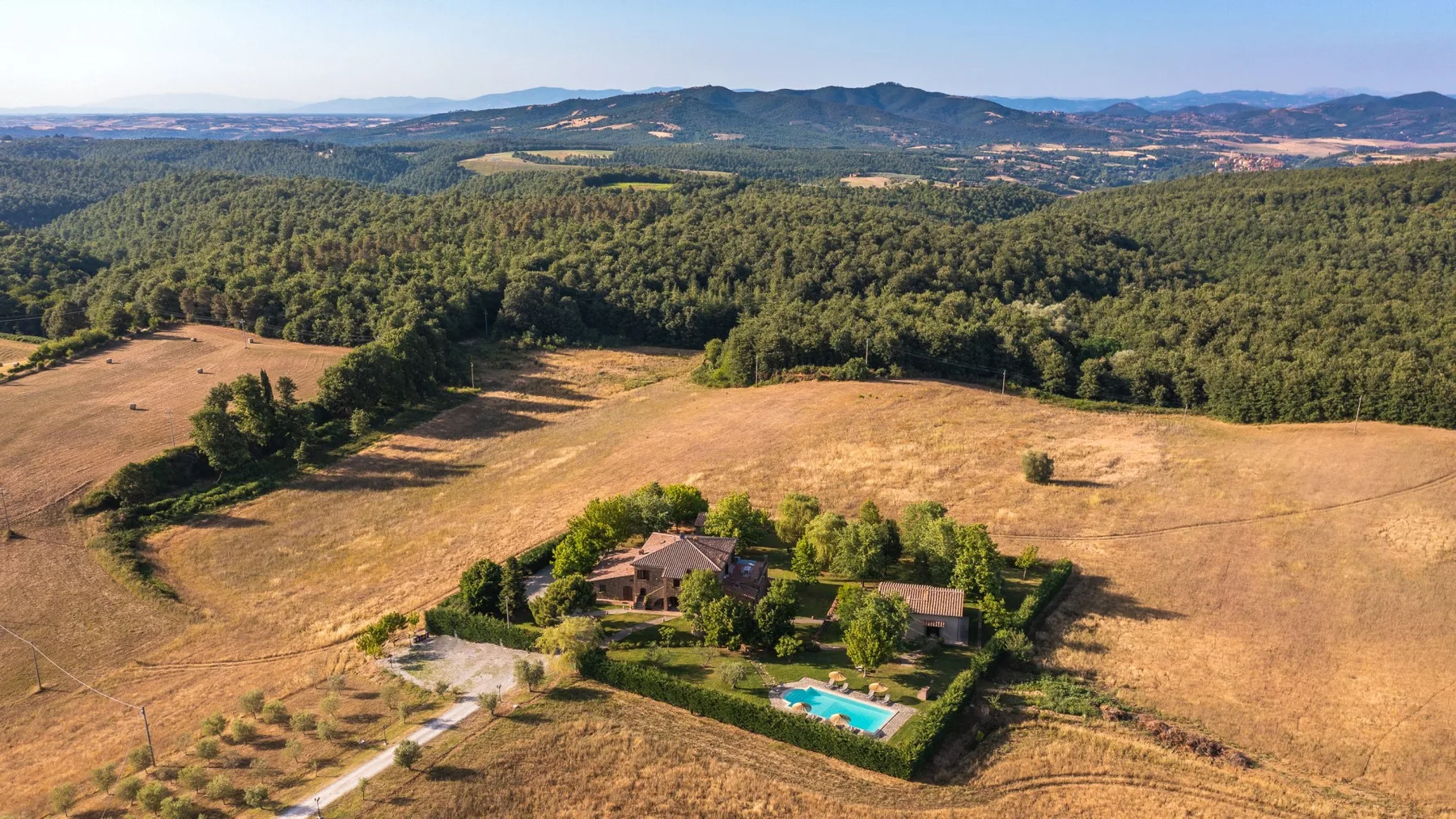 Una vista aérea muestra una casa de campo, un jardín y una piscina entre campos abiertos y colinas boscosas.