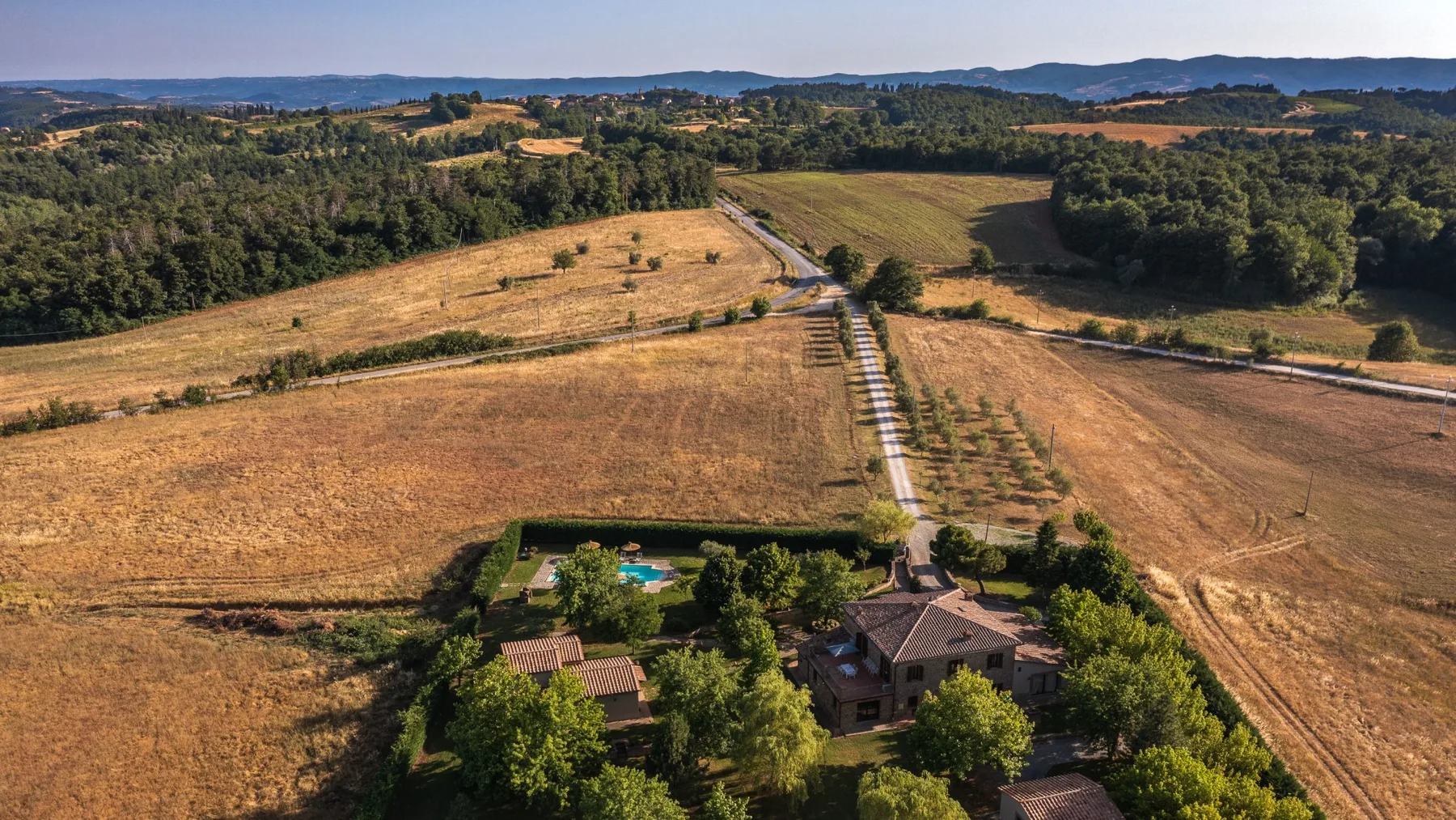 Una vista aérea muestra una propiedad rural rodeada de campos secos, árboles y colinas lejanas.