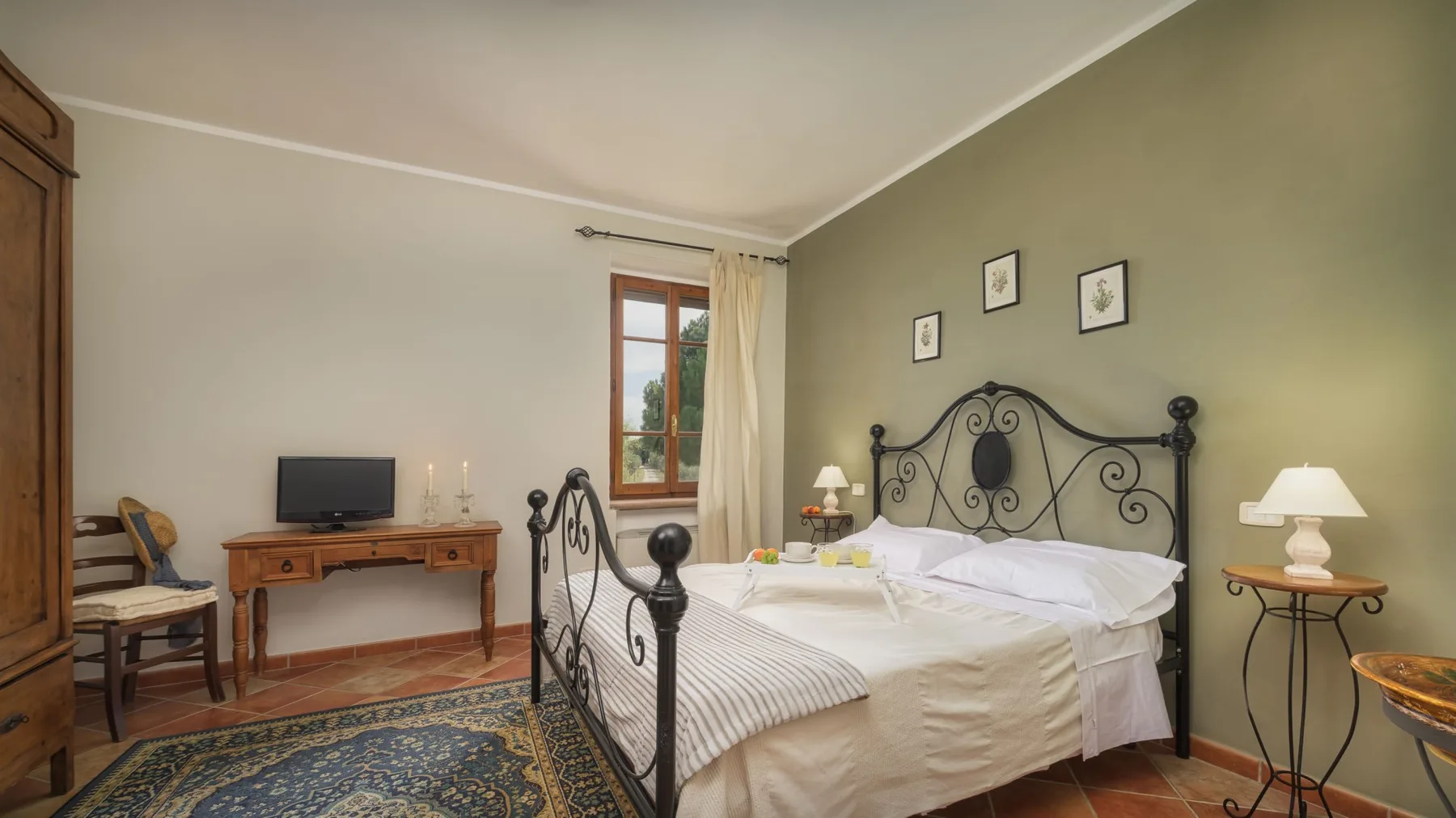 Un dormitorio con cama de hierro, ropa de cama blanca, escritorio de madera y una ventana con vistas a la vegetación.
