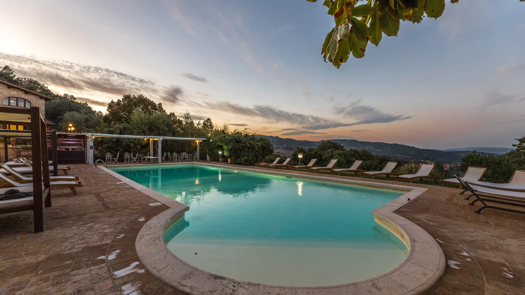 Una piscina al aire libre está junto a una terraza pavimentada, con tumbonas y colinas a lo lejos.