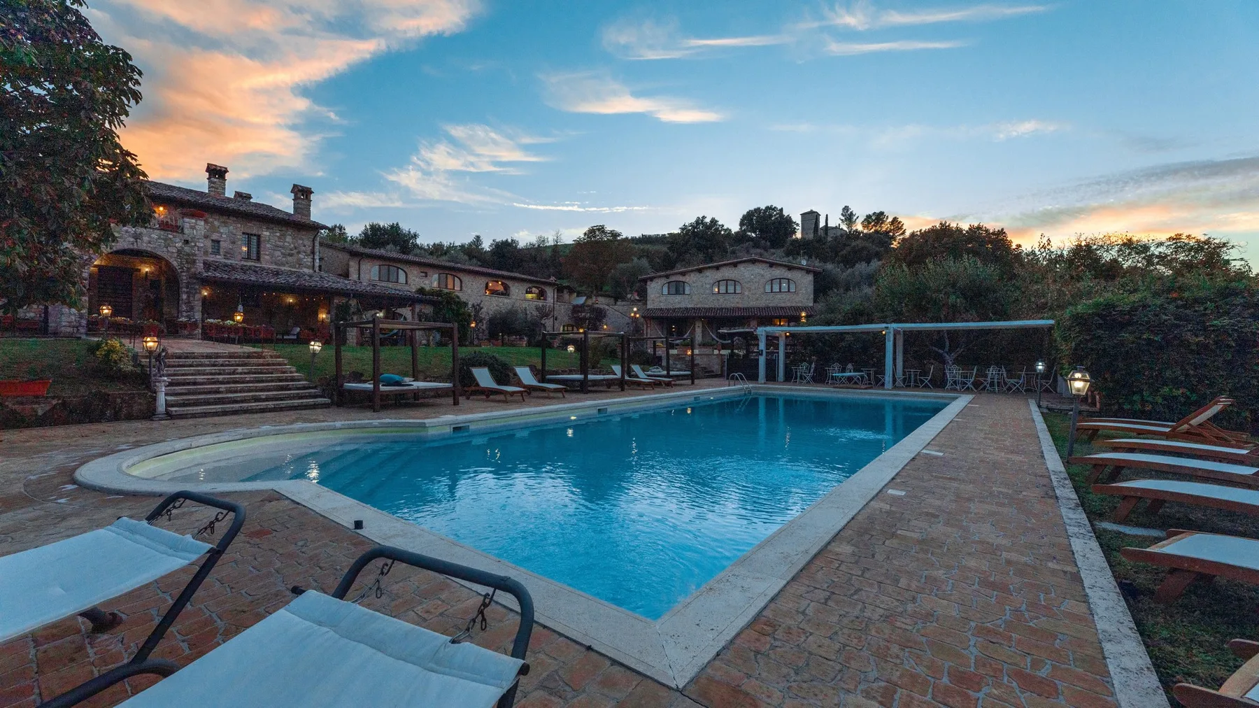 Una piscina al aire libre está rodeada de asientos reclinables y edificios bajos al atardecer.