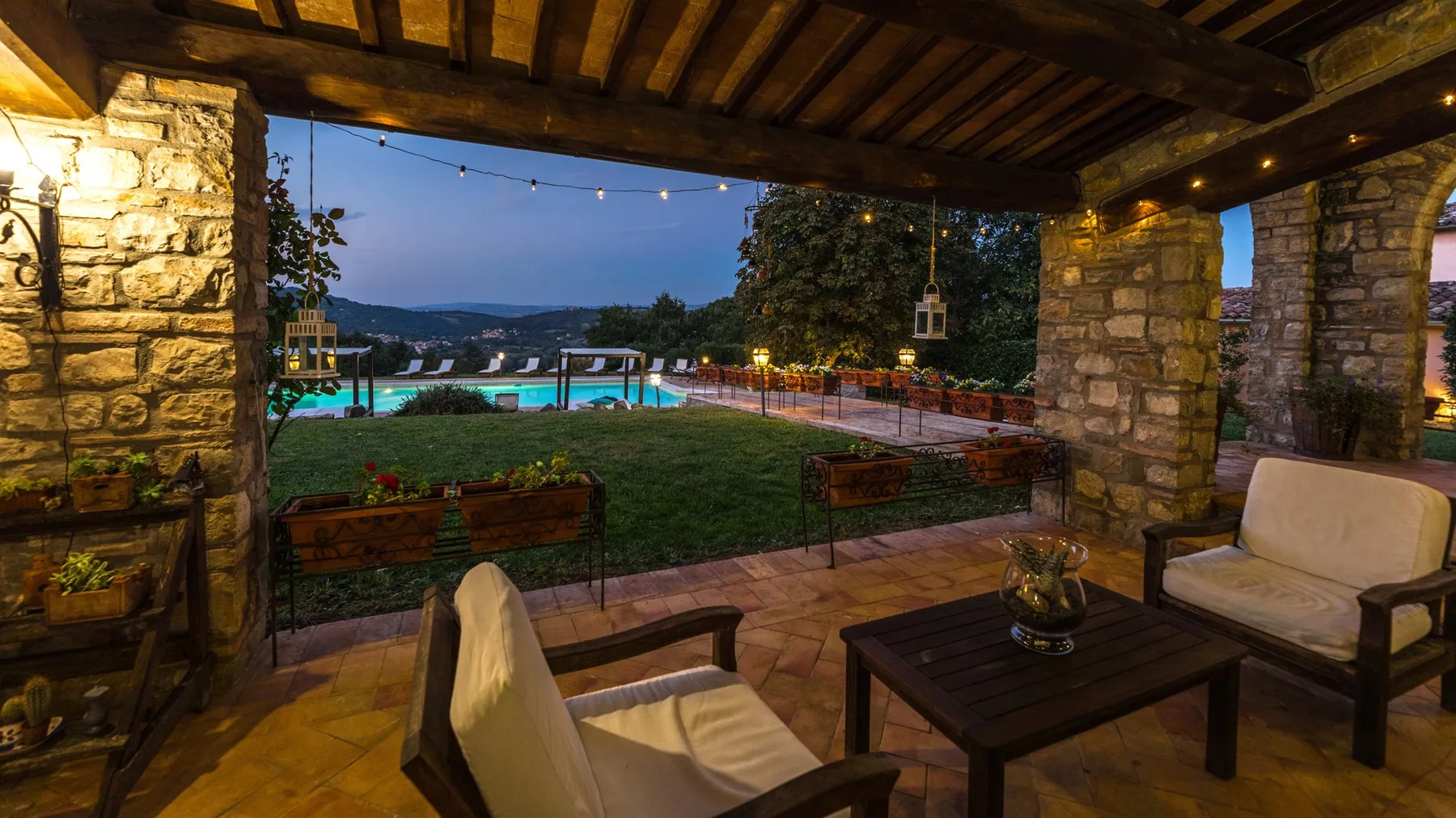 Un patio cubierto de piedra da a un jardín y una piscina iluminada.