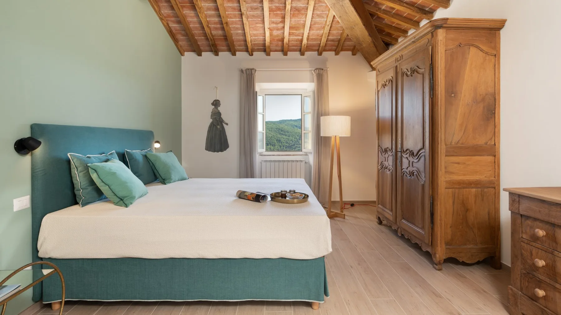 A bedroom with a teal bed, wooden wardrobe, and a window overlooking green hills.