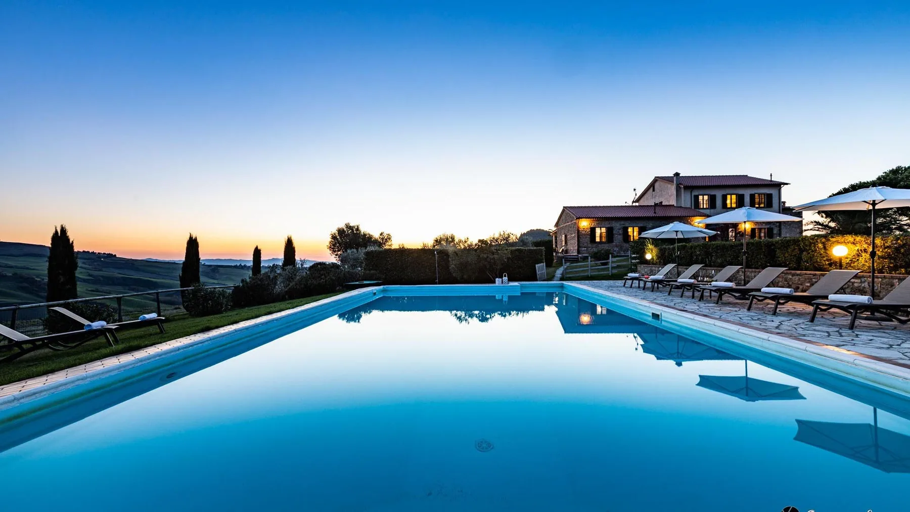 Una piscina al aire libre refleja el cielo al atardecer junto a una casa y asientos.