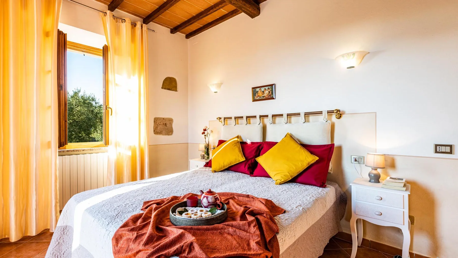 Un dormitorio con una cama grande, cojines coloridos y una ventana iluminada por el sol.