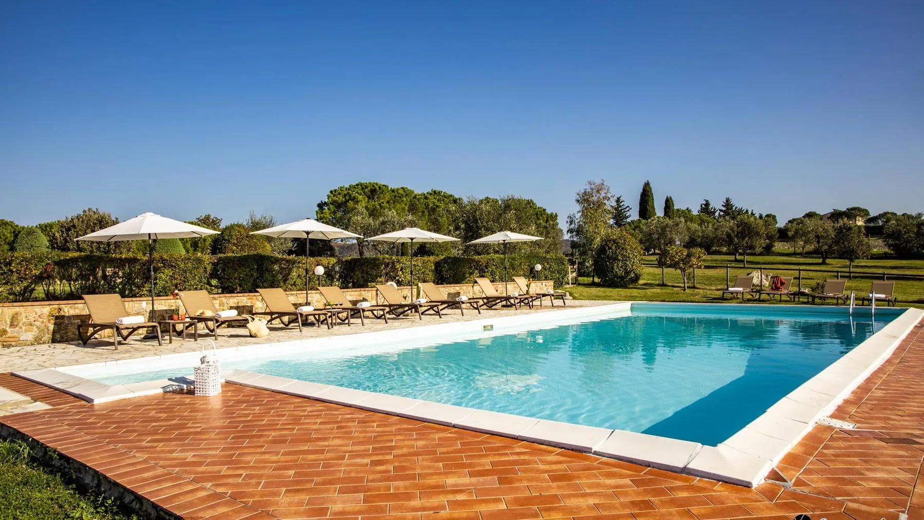 Una piscina al aire libre está rodeada por una terraza embaldosada, asientos y sombrillas.
