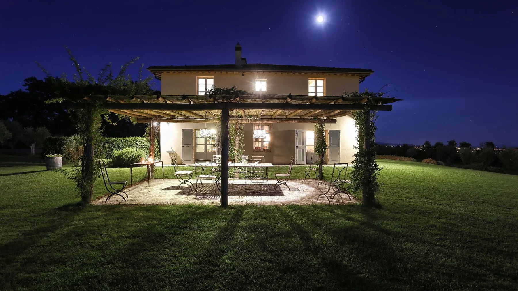 Una casa de dos plantas brilla de noche detrás de una pérgola cubierta de enredaderas, sobre el césped.