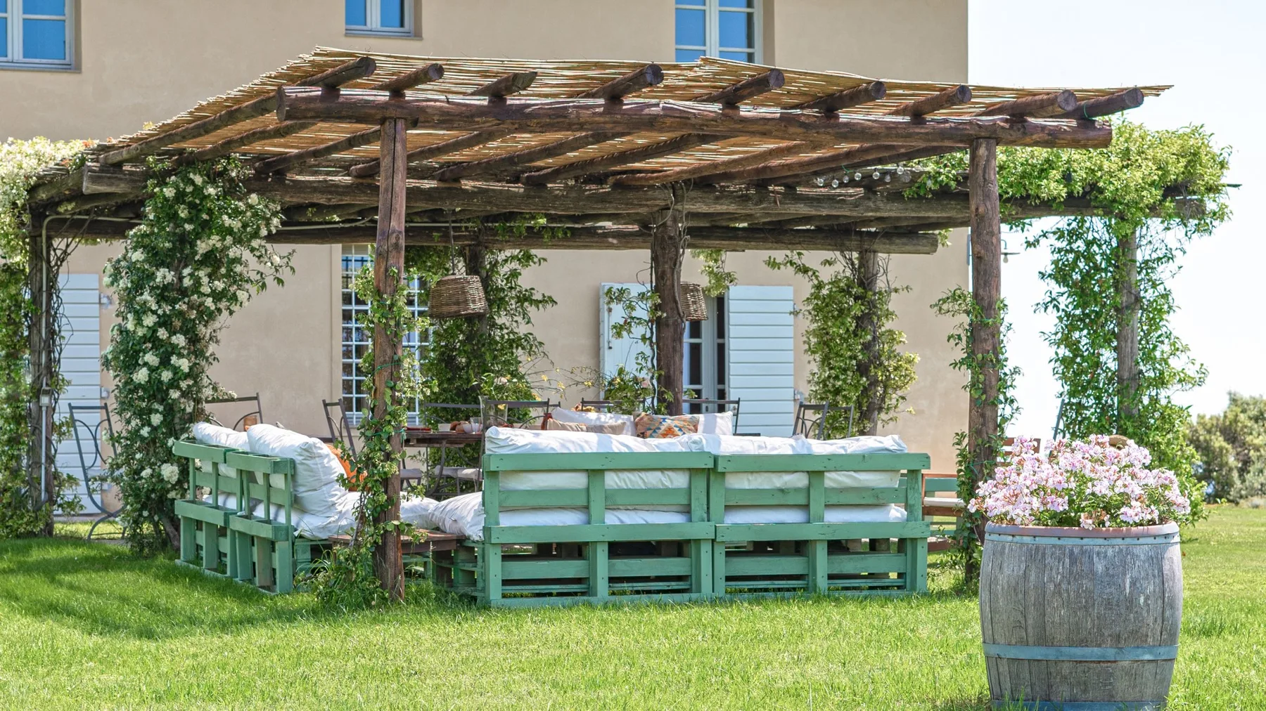 Una zona de estar al aire libre bajo una pérgola de madera, rodeada de vegetación florida.