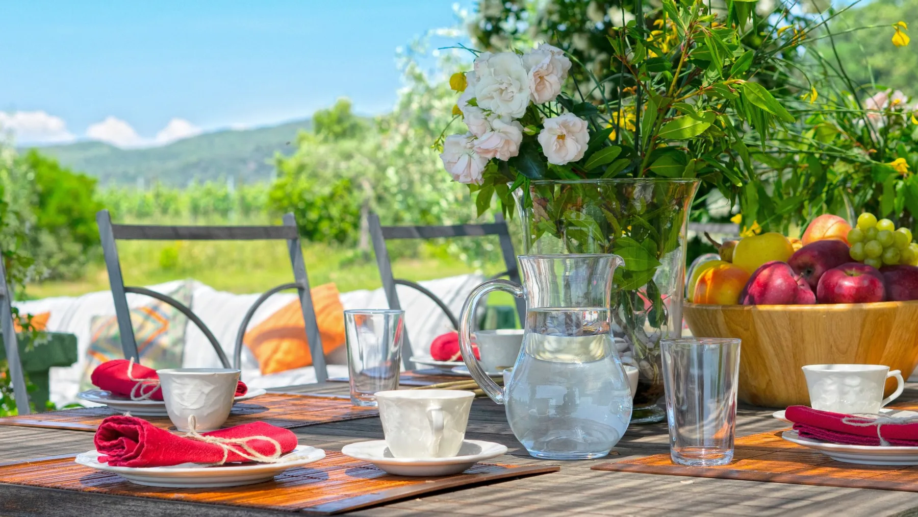 Una mesa de comedor al aire libre está puesta con tazas, una jarra de agua, fruta y flores.