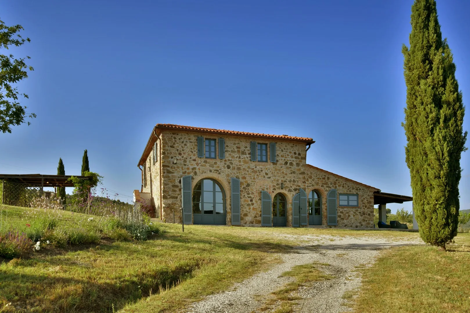 Una casa de piedra con contraventanas verdes está junto a un camino de grava y un ciprés alto.