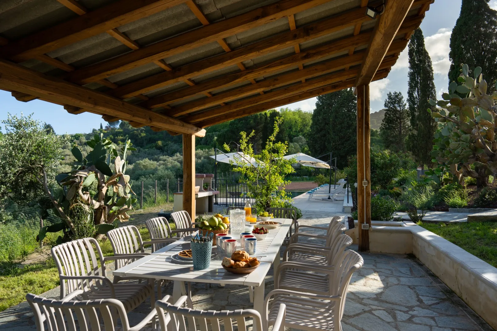 Una zona de comedor exterior cubierta, con una mesa larga y asientos, da a un jardín y una piscina.