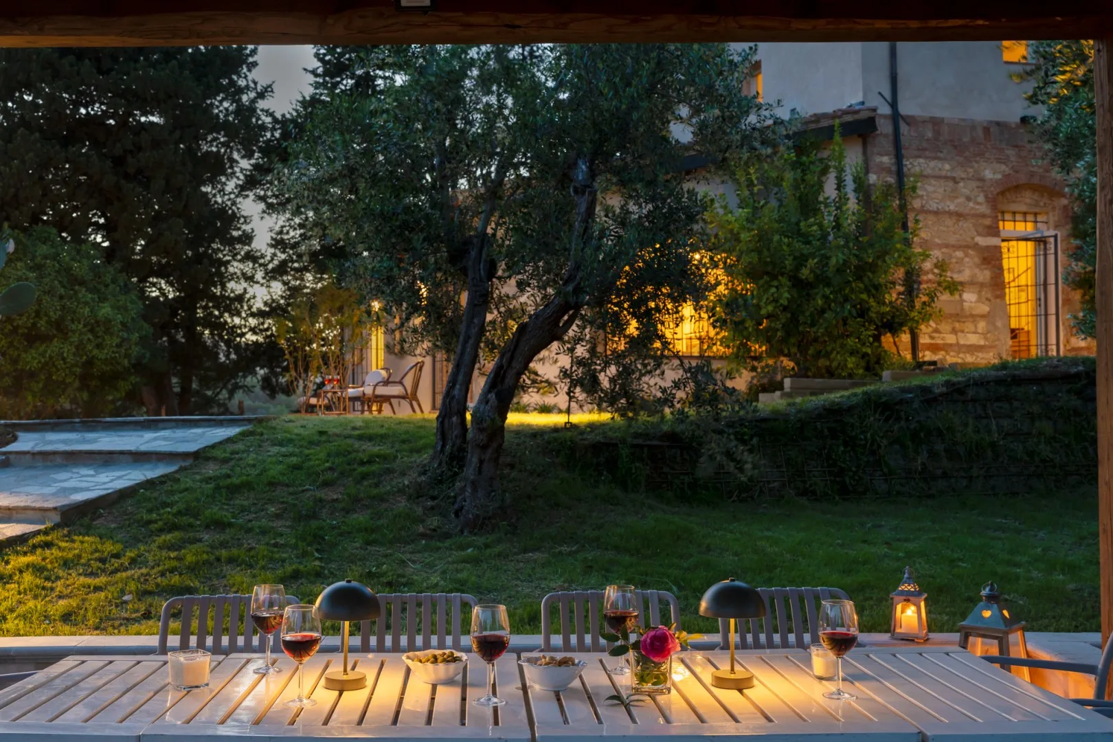Una zona de comedor al aire libre da a un jardín con césped, árboles y un edificio iluminado.
