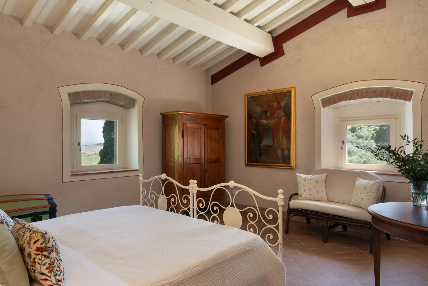 Un dormitorio con una cama, un cabecero de metal ornamentado y asientos bajo dos ventanas.