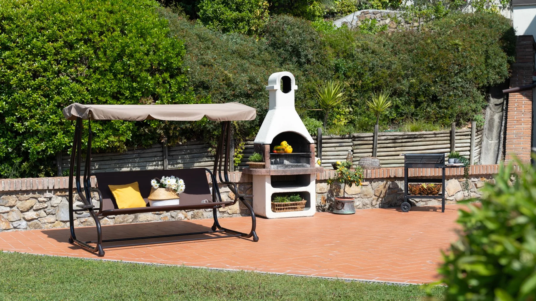 Un patio exterior con un columpio cubierto, una barbacoa y vegetación alrededor.