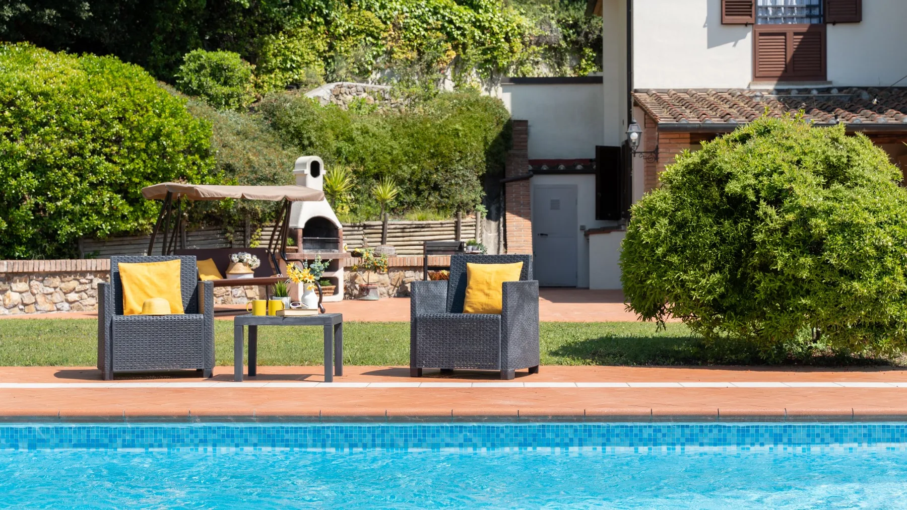 Una piscina exterior junto a un patio con asientos y vegetación alrededor.