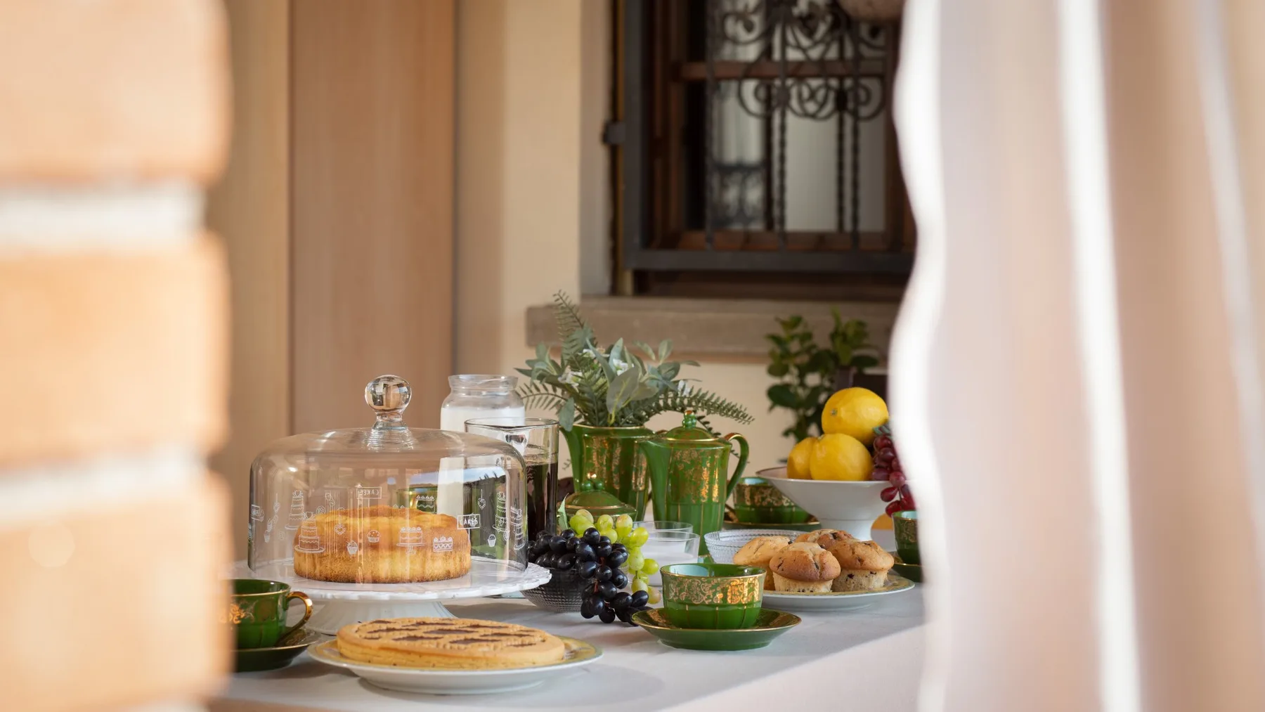 Una mesa de desayuno presenta bollería, fruta y plantas en macetas junto a una ventana.