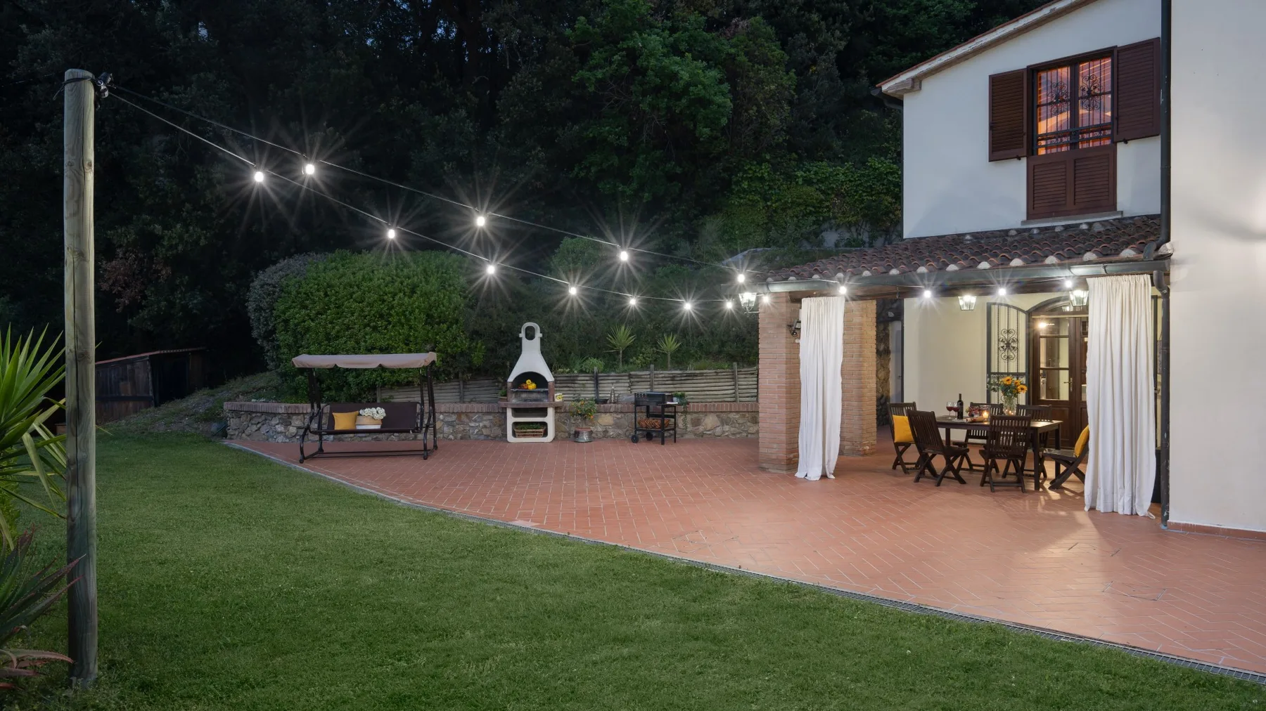 Una zona de patio con asientos junto al césped, iluminada por guirnaldas de luces.