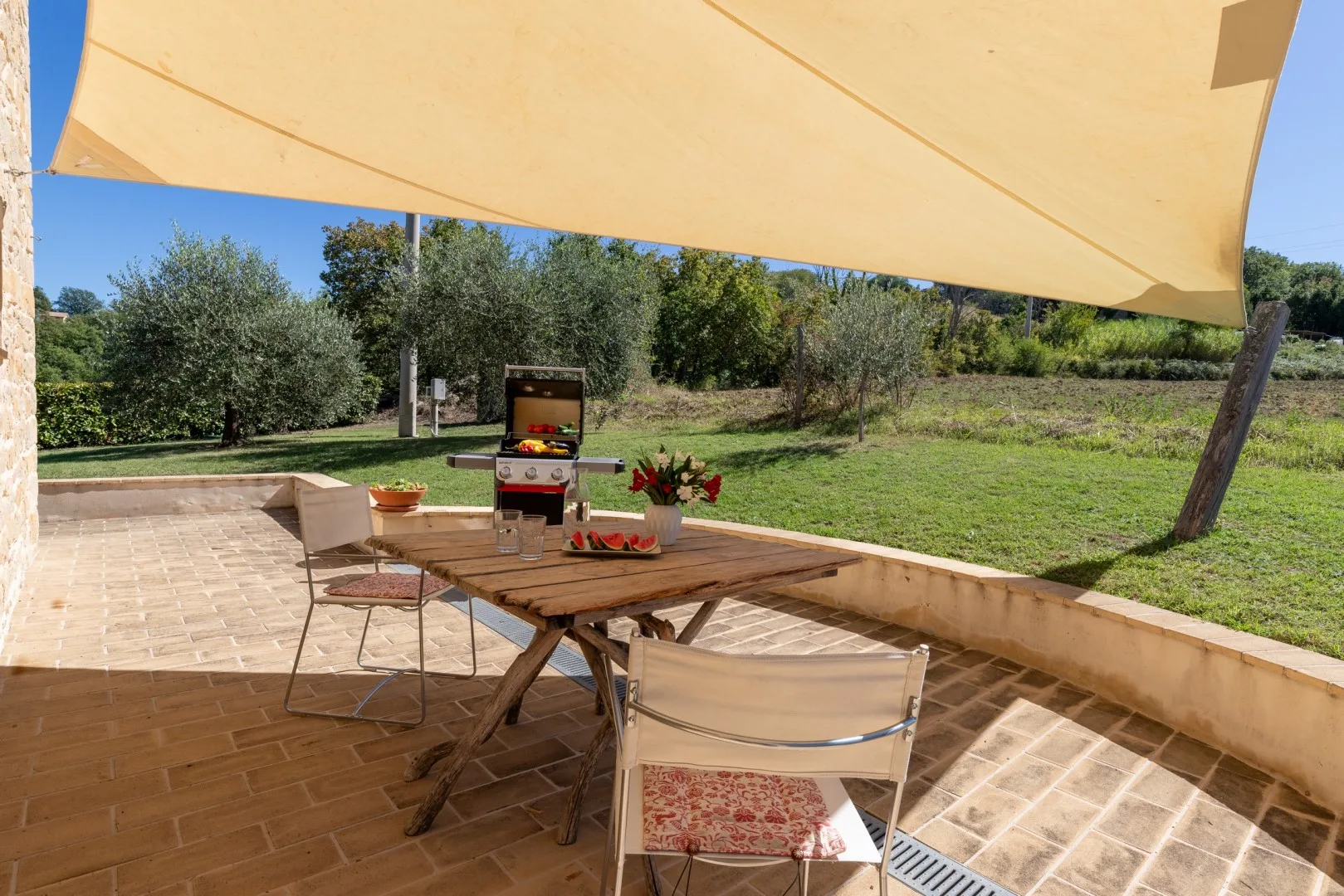 Un patio exterior sombreado tiene una mesa de comedor de madera, sillas, flores en maceta y una barbacoa junto a un jardín con césped.