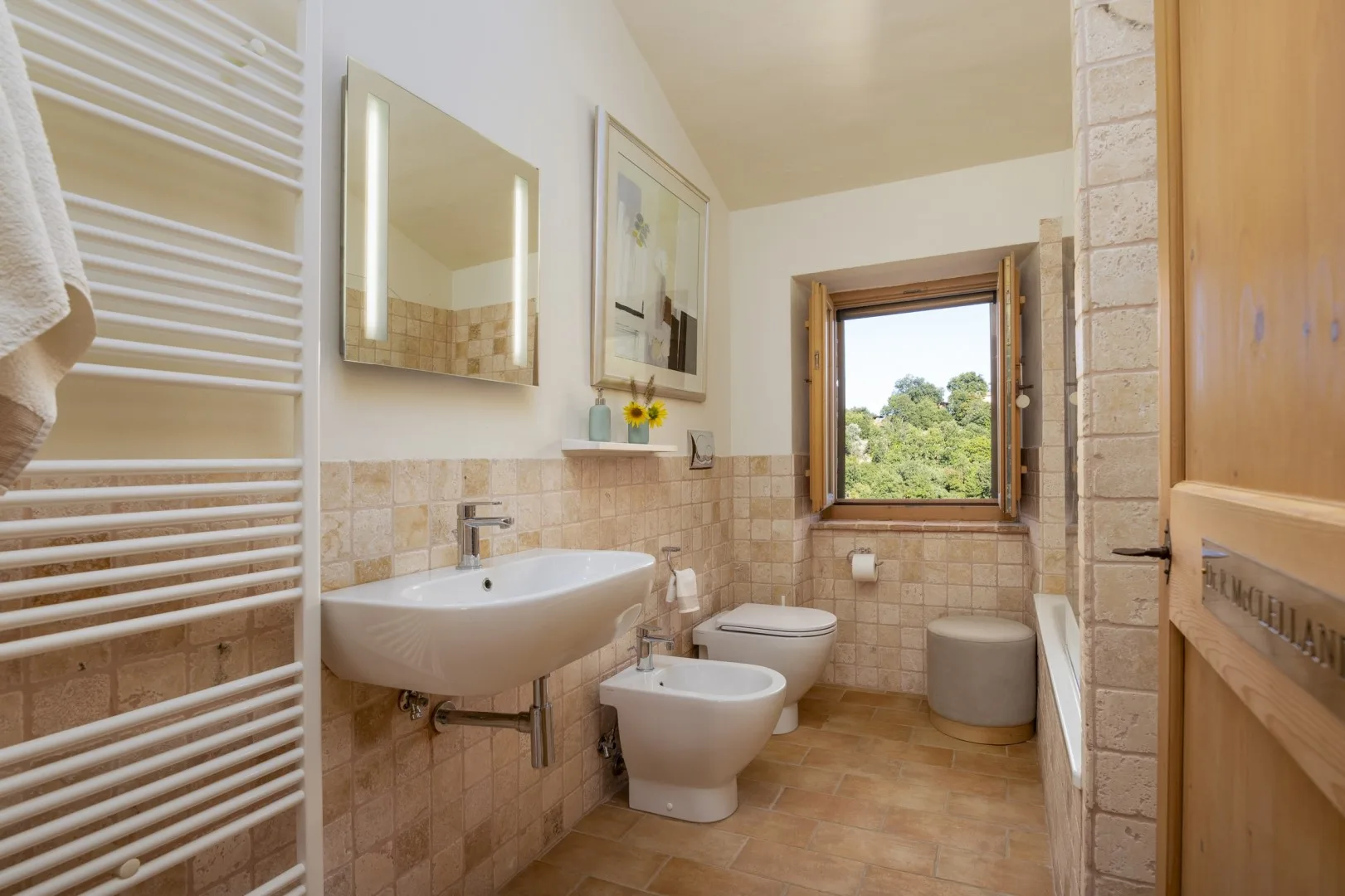 Un baño alicatado tiene un lavabo suspendido, inodoro, bidé y una ventana con vistas a la vegetación.