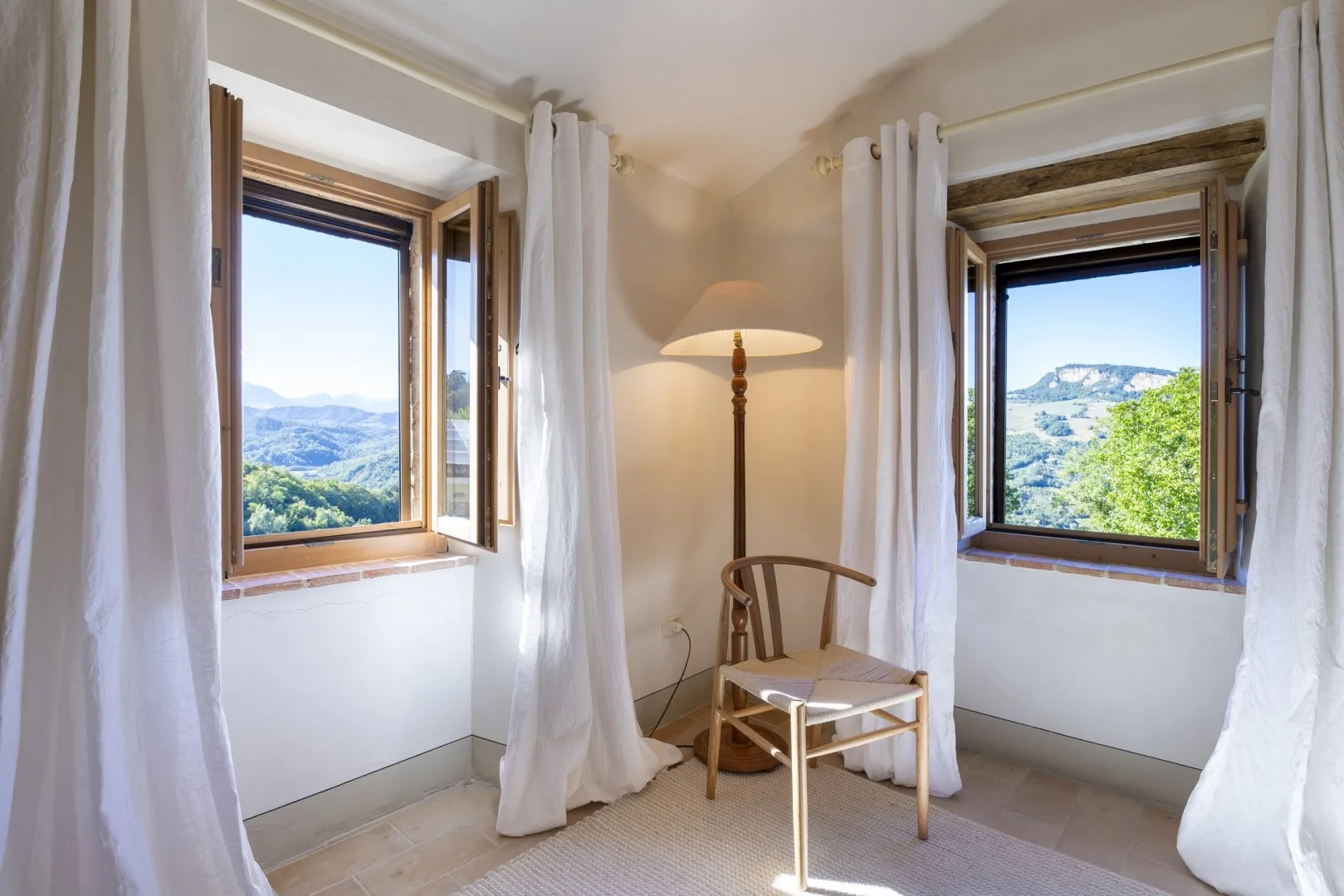 Una habitación luminosa tiene dos ventanas abiertas con cortinas blancas, una lámpara de pie, una silla y una mesita.