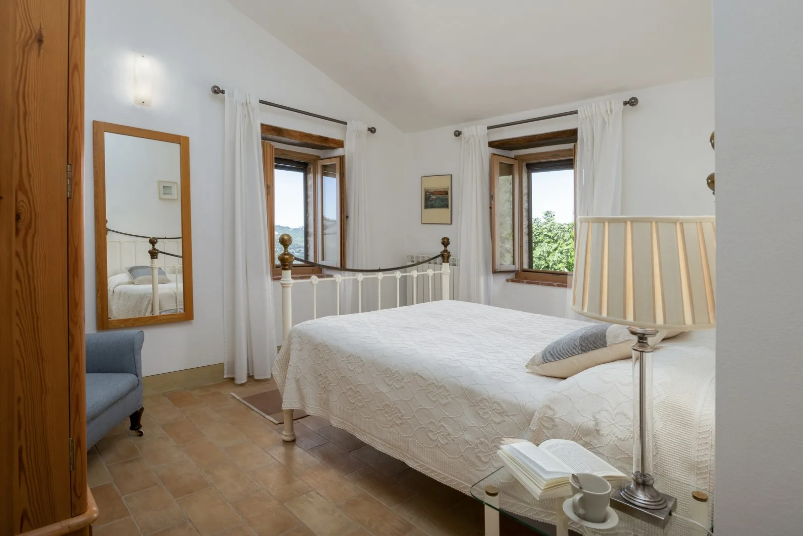 Un dormitorio luminoso con una cama blanca, dos ventanas con cortinas y un pequeño sillón azul.