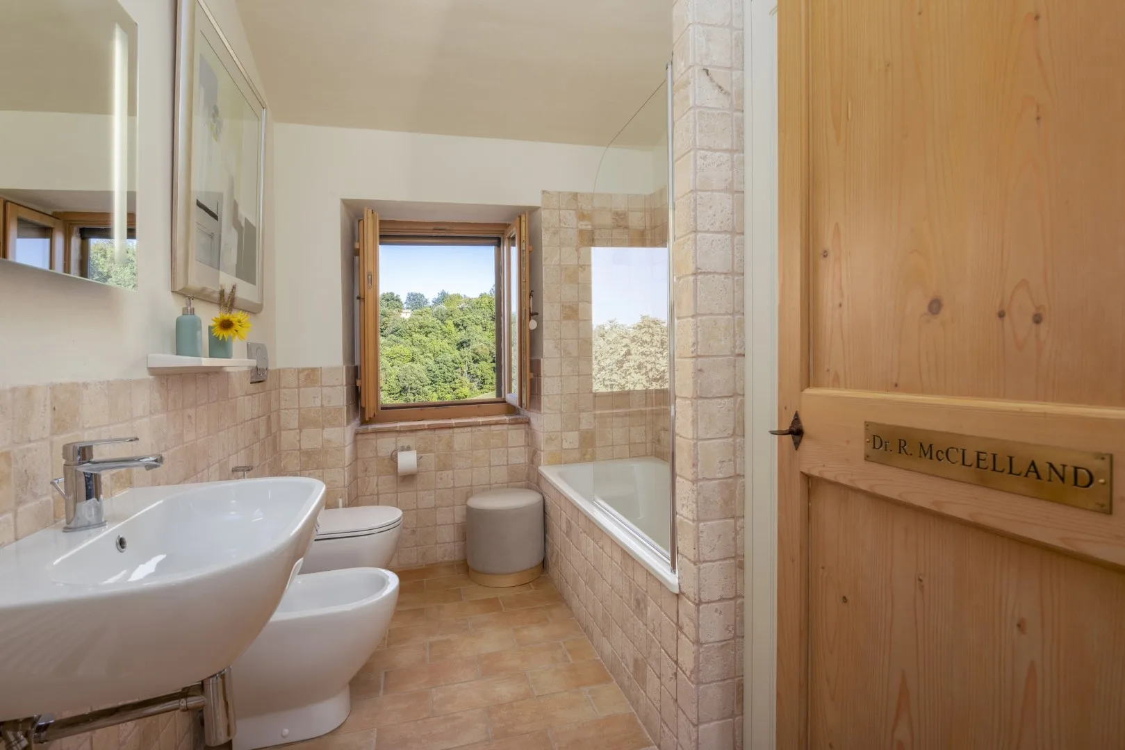 Un baño alicatado tiene un lavabo y un inodoro blancos, una bañera y una ventana con vistas a árboles verdes.