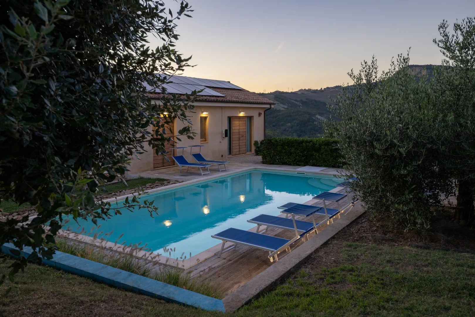 Una piscina turquesa con tumbonas está junto a una casa pequeña al atardecer, con colinas al fondo.
