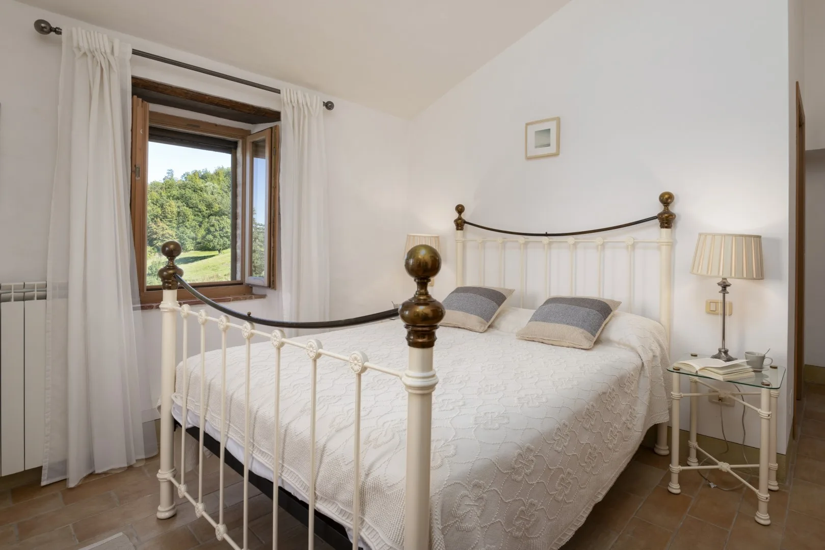 Un dormitorio luminoso con una cama de metal blanco, ropa de cama estampada y una ventana con vista a árboles frondosos.
