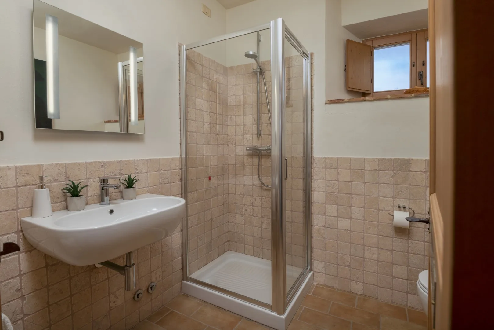 Un baño alicatado con ducha de vidrio, lavabo suspendido, espejo y una ventana pequeña.