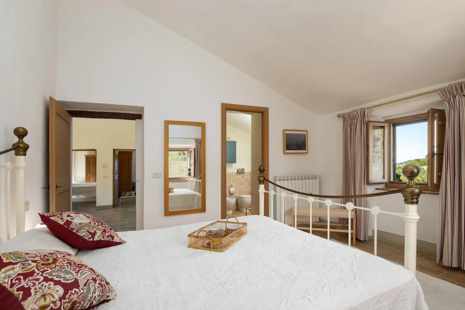 Un dormitorio luminoso tiene una cama grande blanca, cojines estampados, una mesita de noche y una ventana abierta.