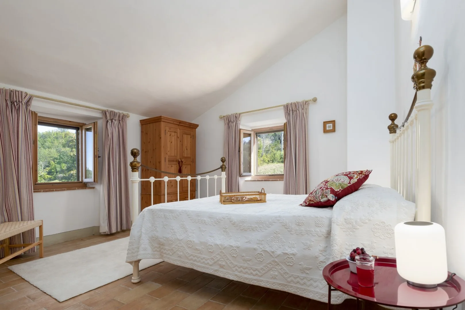 Un dormitorio luminoso cuenta con una cama blanca, un armario de madera, dos ventanas con cortinas y una mesita roja.