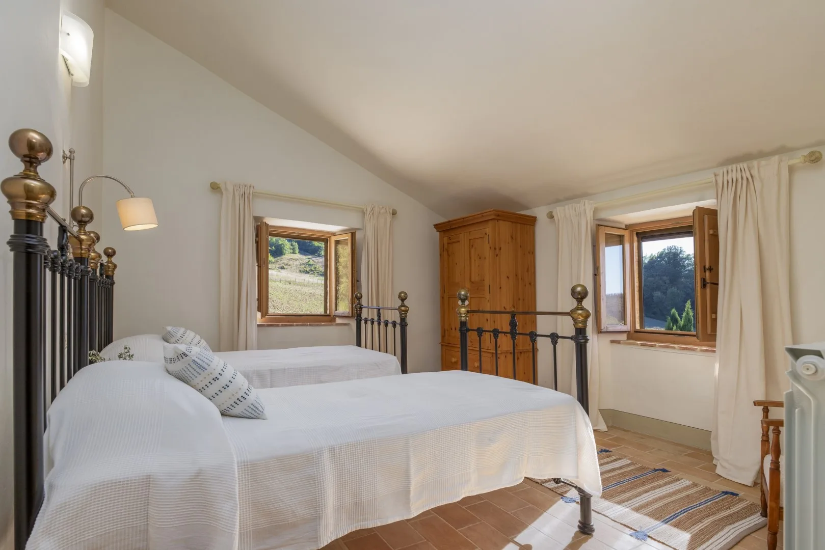 Un dormitorio con dos camas blancas, un armario de madera y dos ventanas con cortinas.