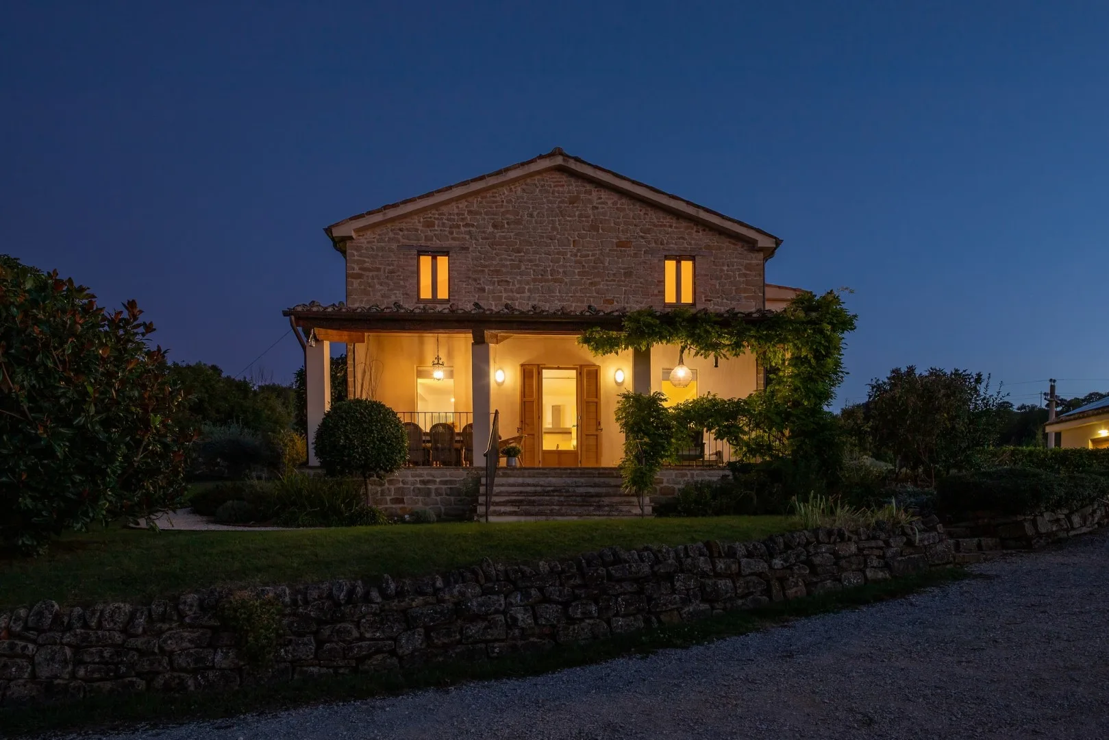Una casa de piedra brilla al anochecer, con un porche iluminado, ventanas superiores y un muro bajo de jardín.