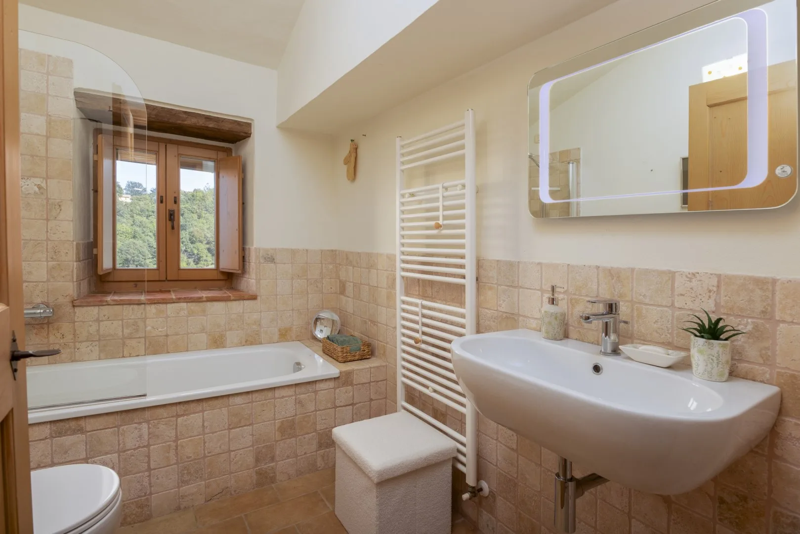 Un baño alicatado con bañera bajo una ventana, lavabo mural y toallero calefactable.