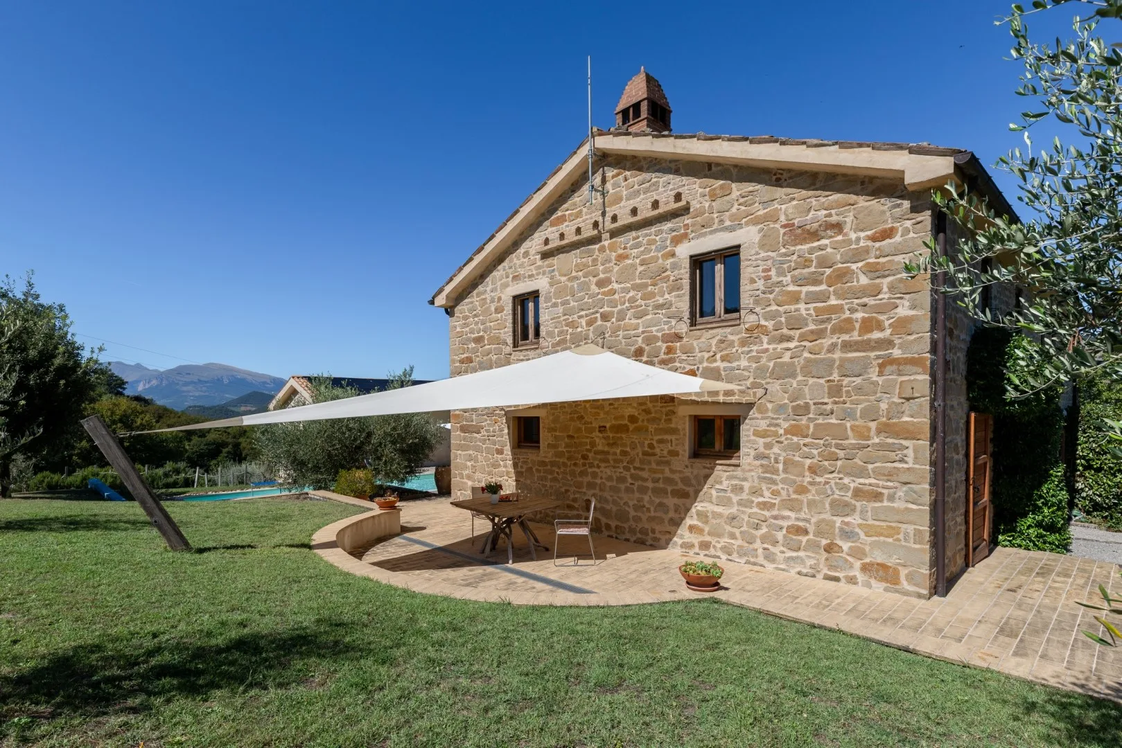 Una casa de piedra da a un jardín con césped y un patio sombreado bajo una cubierta blanca.