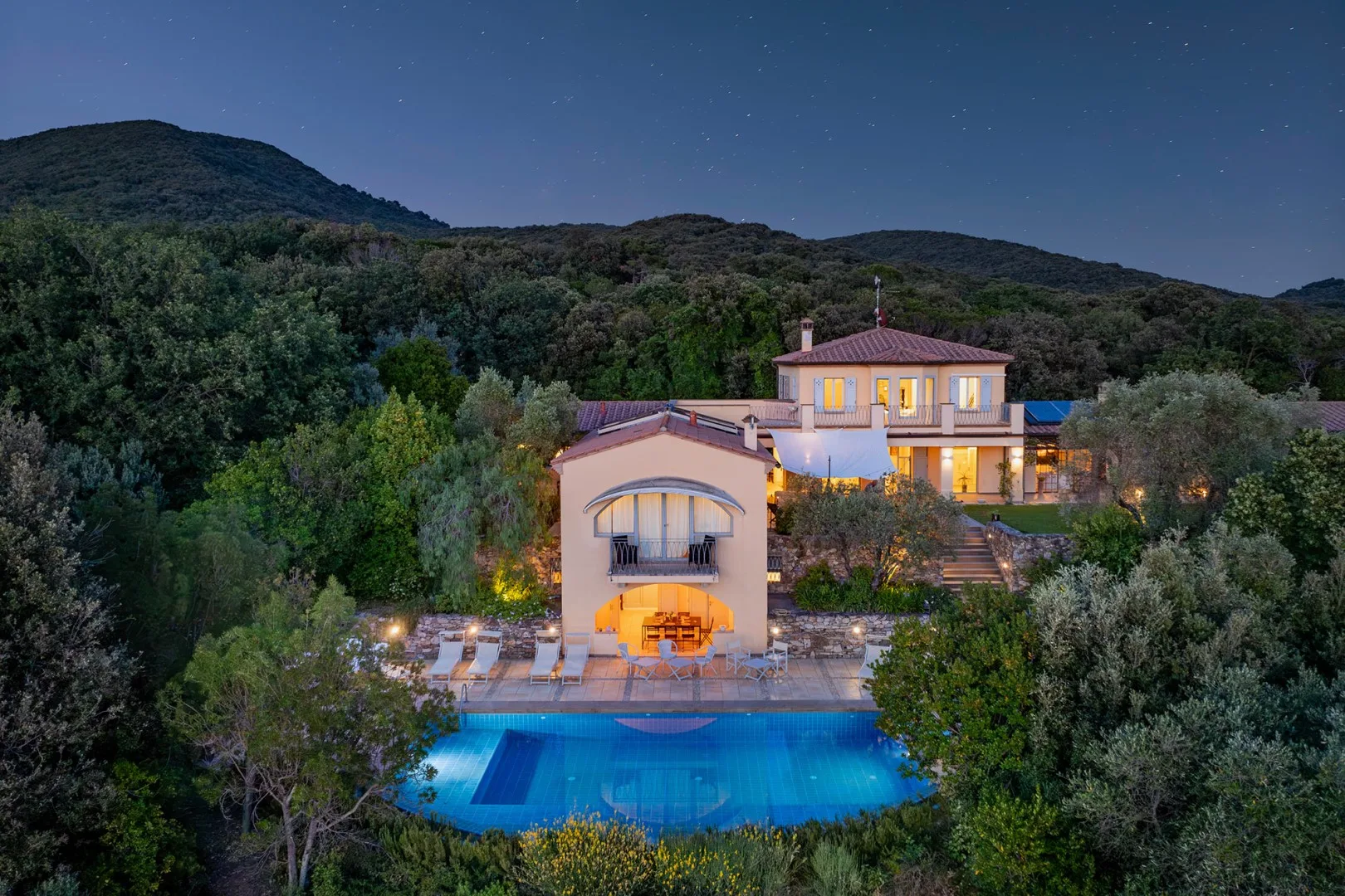 Una villa en la ladera brilla al anochecer sobre una piscina, rodeada de colinas boscosas.