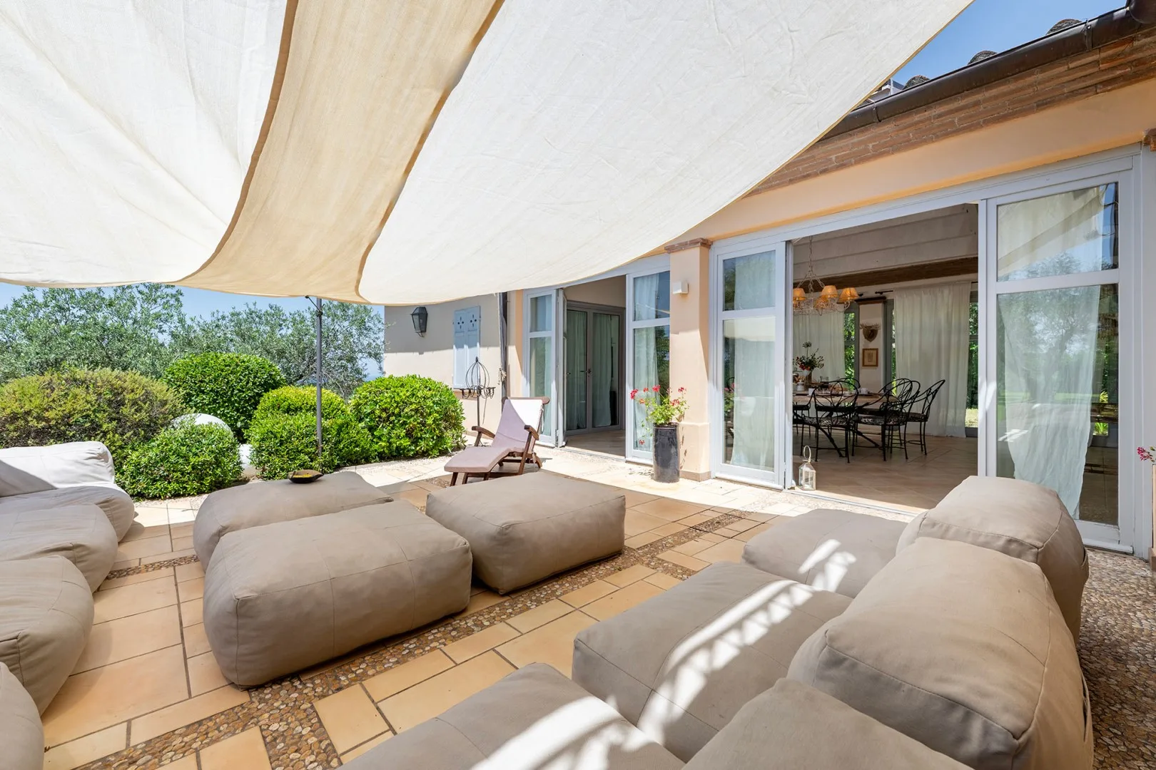 Un patio exterior a la sombra, con asientos acolchados y puertas abiertas a un comedor.