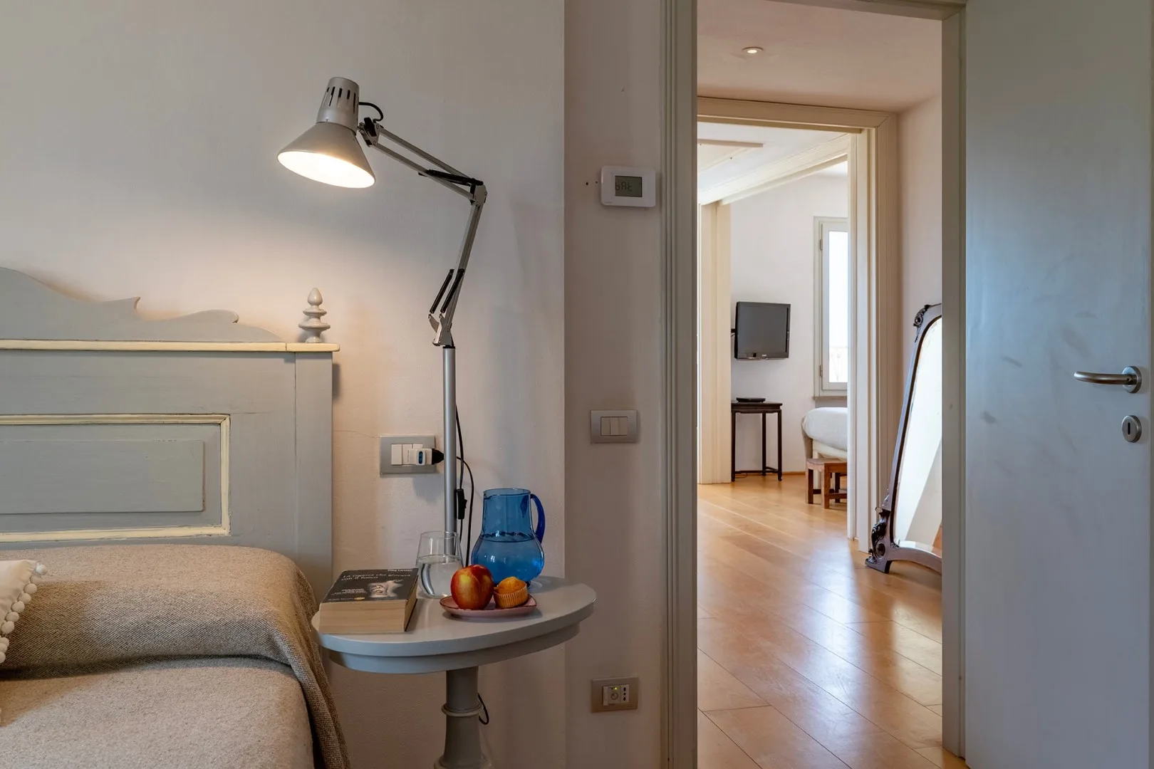 Un dormitorio con una mesa auxiliar y una lámpara, junto a una puerta que da a una habitación luminosa.