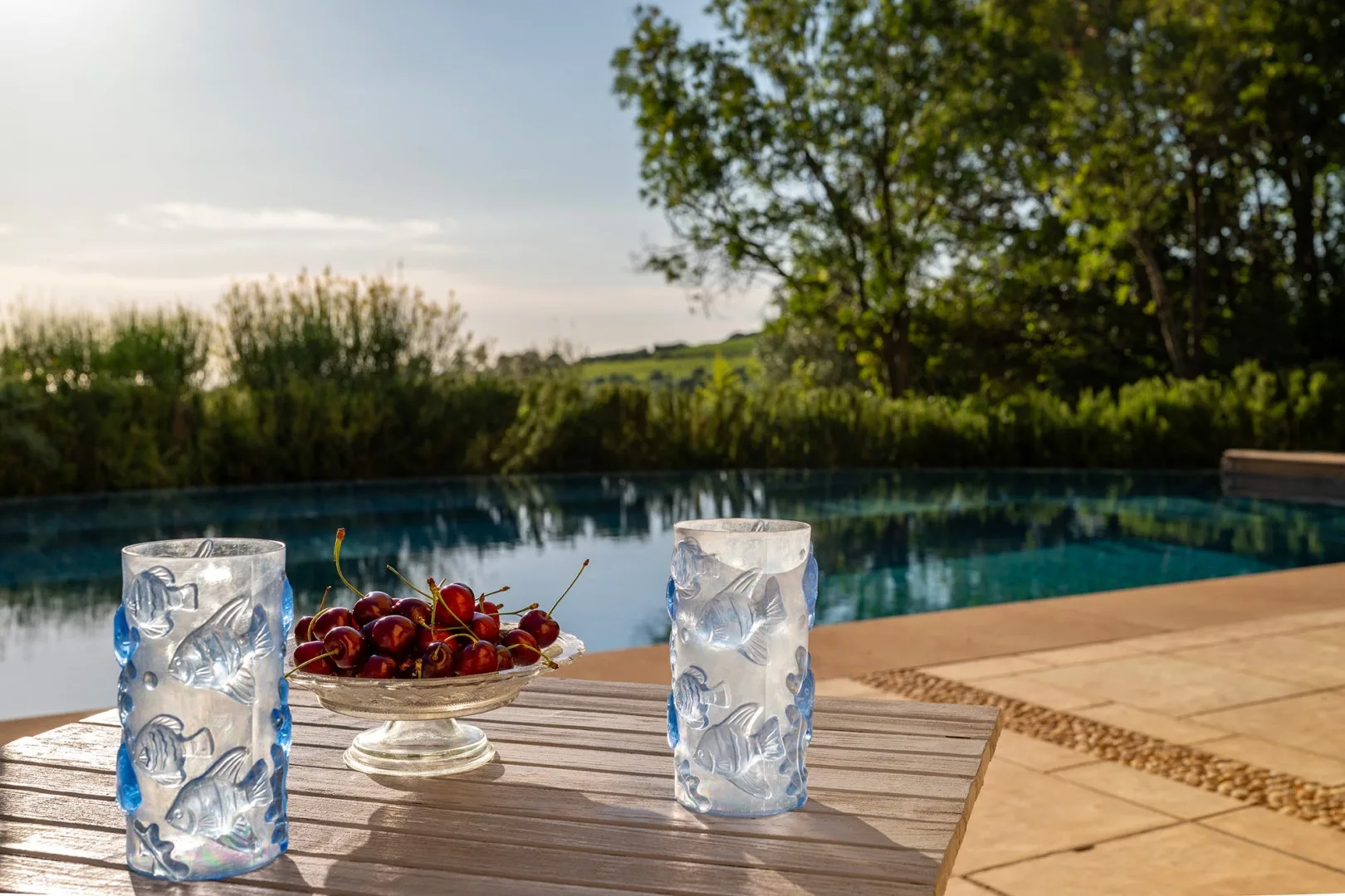 Dos vasos y un cuenco de cerezas están junto a una piscina.